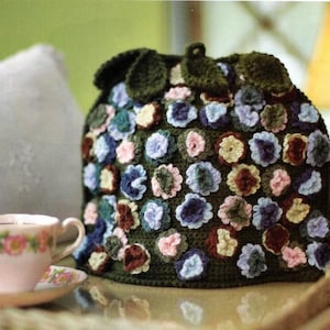 Puede incluir: Un cubrete de té de ganchillo hecho a mano, verde oliva, adornado con aplicaciones florales tridimensionales de colores. El cubrete tiene una parte superior en forma de hoja y un lazo. Una taza de té y un platillo con motivos florales están en primer plano.