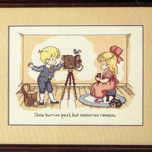 May include: Cross-stitch framed artwork depicting two children with a vintage camera. The boy is adjusting the camera on a tripod, while the girl sits nearby. The artwork includes the text "Time hurries past, but memories remain."