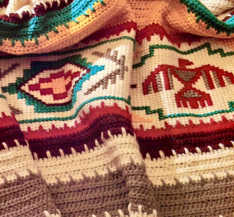 Puede incluir: Una manta de ganchillo con un dise&ntilde;o del suroeste que presenta un p&aacute;jaro, formas geom&eacute;tricas y rayas en tonos de rojo, verde, marr&oacute;n y blanco.