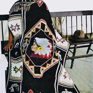 Pode incluir: Uma manta de crochê preta com um design nativo americano colorido que apresenta um perfil de contas de uma pessoa usando um cocar. A manta tem franjas na parte inferior e está drapeada sobre um banco de madeira.
