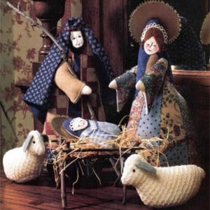 May include: A handmade nativity scene featuring three figures: Mary, Joseph, and baby Jesus. The figures are dressed in colourful patchwork fabric and have painted faces. Two white lambs are also included in the scene.