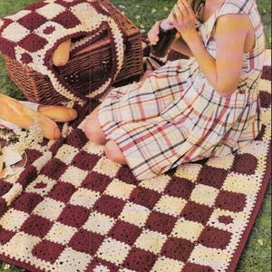 Peut inclure: Une couverture en crochet rectangulaire présentant un motif en damier composé de carrés alternés de couleur bordeaux foncé et crème. La couverture est réalisée avec une finition en fil texturé, affichant un motif en grille cohérent.