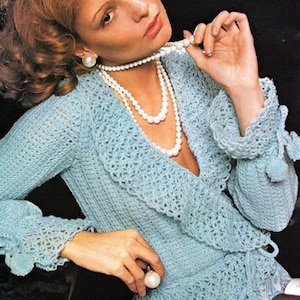 Könnte beinhalten: Ein hellblauer Häkelcardigan mit Rüschenkragen und langen Ärmeln. Der Cardigan ist mit einer Schärpe in der Taille gebunden und hat Bommeln an den Ärmeln. Das Model trägt eine Perlenkette und Ohrringe.