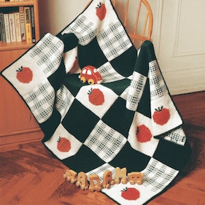 Puede incluir: Una manta hecha a mano, a cuadros, con cuadrados verdes oscuros y blancos alternados. Cada cuadrado blanco presenta un diseño de manzana roja. Un pequeño coche de juguete rojo y un juego de trenes de madera están sobre la manta, que está sobre una silla de madera.