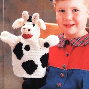 May include: A white crocheted cow puppet with black spots. The puppet has a red mouth and a black nose. A child is holding the puppet.