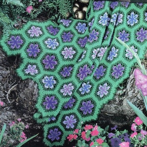 Pode incluir: Uma manta de crochê com fundo verde e flores roxas e azuis. A manta é feita de um fio macio e texturizado e tem um design delicado e feminino.