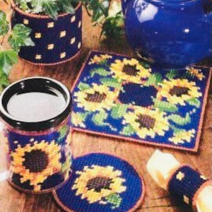 May include: A set of four sunflower-themed crocheted coasters and a matching mug cozy. The coasters are round and square, with a blue background and yellow sunflowers. The mug cozy is cylindrical and features a single sunflower.