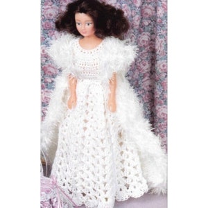 Può includere: Un abito bianco all'uncinetto con una gonna lunga e una giacca bolero abbinata. L'abito è decorato con un delicato bordo in pizzo. La bambola indossa l'abito e ha lunghi capelli castani.