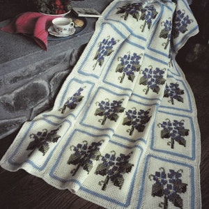 May include: A crocheted throw blanket with a white background and blue and green floral pattern. The blanket is folded and draped over a stone surface.