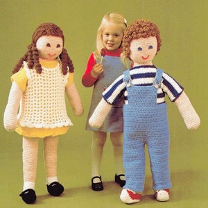 May include: Two crocheted dolls, one girl and one boy, stand next to a young girl holding a lollipop. The girl doll wears a white crocheted dress with a yellow underskirt and brown curly hair. The boy doll wears blue overalls with a white and blue striped shirt and brown curly hair.