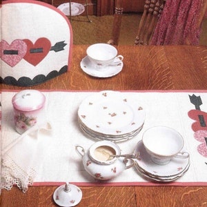 May include: A Valentine's Day themed tea set with a white tea cozy, a table runner, and a tea service. The tea cozy and table runner feature heart and arrow designs. The tea set includes cups, saucers, plates, a sugar bowl, and a creamer.