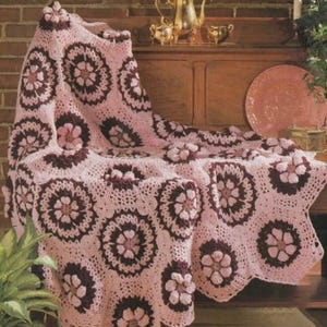 May include: A handmade crochet blanket featuring a repeating floral motif pattern in light pink and dark brown yarn. The textile is constructed from joined circular and square segments, creating a textured, scalloped edge throughout the design.