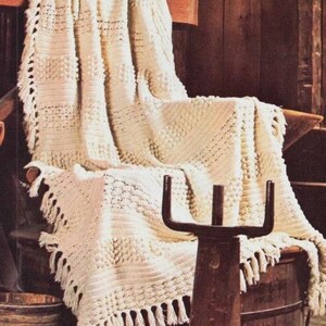 May include: Cream-coloured, hand-crocheted blanket with a textured pattern and fringe detail. The blanket is draped over a wooden chair and barrel, creating a cosy, rustic aesthetic. The blanket's dimensions are not visible.
