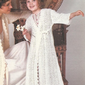 May include: A young girl wearing a white crocheted robe with a tie belt. She is holding a white daisy in her hand. The robe has a lacy trim around the edges.