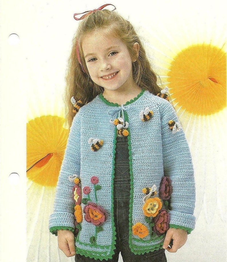 Puede incluir: Un c&aacute;rdigan de punto azul claro con ribetes verdes y aplicaciones de abejas y flores. El c&aacute;rdigan lo lleva una ni&ntilde;a peque&ntilde;a con el pelo largo y casta&ntilde;o.