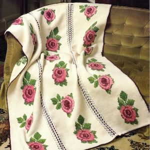 May include: A white crocheted throw blanket with a repeating pattern of pink roses and green leaves. The blanket is draped over a brown couch.