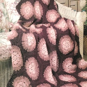 May include: A crochet blanket featuring a repeating pattern of hexagonal motifs. Each motif consists of a dark brown outer border surrounding a circular center that transitions from a light pink to a soft mauve hue, creating a floral-like appearance.
