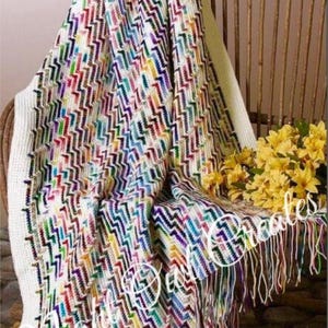 Peut inclure: Une couverture en crochet colorée avec un fond blanc et un motif en chevron. La couverture a des franges sur les bords et est drapé sur une chaise en bois.