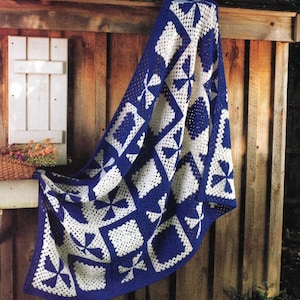 Può includere: Una coperta all'uncinetto blu e bianca con un motivo geometrico. La coperta è fatta di quadrati con diversi disegni, tra cui girandole e cerchi. La coperta è appesa a una recinzione di legno.