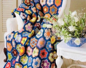 Op de afbeelding: Een gehaakt deken met een blauwe achtergrond en kleurrijke bloemmotieven. Het deken is gedrapeerd over een witte fauteuil met een vaas met witte bloemen op een witte tafel ernaast.