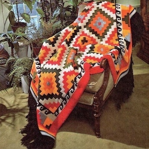 May include: A colorful crocheted throw blanket with geometric patterns in shades of orange, red, green, black, and white. The blanket is draped over a wooden armchair with a fringed edge.
