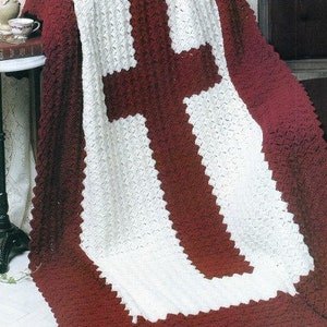 May include: A crocheted throw blanket with a large white cross on a burgundy background. The blanket is made with a textured stitch and has a fringed edge.