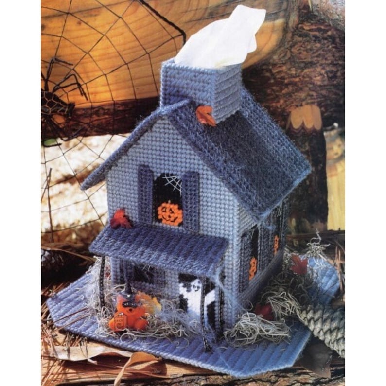 May include: A blue and gray Halloween-themed tissue box cover shaped like a house with a chimney, a porch, and a window with a jack-o-lantern inside. The house is decorated with a spider web, a ghost, and a black cat.