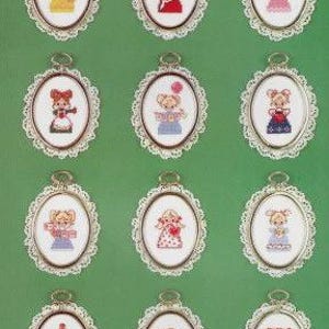 May include: A collection of twelve framed cross-stitch ornaments. Each oval frame has a silver-colored metal edge and a white lace border. The cross-stitch designs feature various figures in colorful outfits, with blonde hair and rosy cheeks, on a green background.