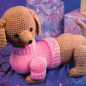 May include: A crocheted dachshund wearing a pink sweater. The dachshund is brown and has a black nose and eyes. The dachshund is sitting on a blue surface.