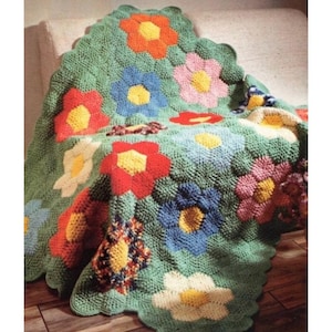 Op de afbeelding: Een gehaakte deken met een bloemenpatroon. De deken is groen met bloemen in rood, oranje, blauw, roze en wit. De bloemen hebben gele centra en zijn in een herhalend patroon gerangschikt. Met een geschulpte rand.
