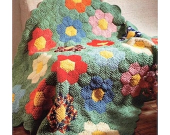 Op de afbeelding: Een gehaakte deken met een bloemenpatroon. De deken is groen met bloemen in rood, oranje, blauw, roze en wit. De bloemen hebben gele centra en zijn in een herhalend patroon gerangschikt. Met een geschulpte rand.