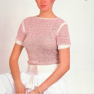 May include: A light pink crochet top with short sleeves and a round neckline. The top has white trim on the sleeves and neckline. A white drawstring is tied at the waist. The top is paired with a white skirt.