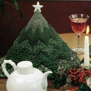 Könnte beinhalten: Ein gesteppter Weihnachtsbaum-Tischaufsatz in Grüntönen mit einem weißen Stern. Der Baum ist mit weißen Blumenmustern verziert. Eine weiße Teekanne, Kerzen und ein Glas mit roter Flüssigkeit sind ebenfalls zu sehen.