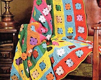 Op de afbeelding: Een kleurrijk gehaakt deken met een bloemenpatroon. Het deken is gemaakt met strepen in verschillende kleuren, waaronder rood, oranje, geel, groen, blauw en roze. Het deken is over een stoel gelegd.