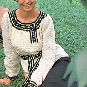 May include: A woman wearing a white crocheted sweater with black and white trim. The sweater has a belt and a V-neck. She is wearing a black skirt and holding a bouquet of pink and red flowers.