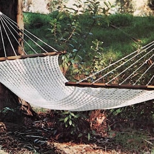 以下が含まれることがあります： 自然な屋外環境にある2本の木の間につるされた白い織りのハンモック。ハンモックは木製のサポートバーがあり、白いロープで張られています。背景には緑の葉と日光が写っています。