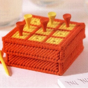 May include: A handmade orange and yellow tic-tac-toe game. The game board is a square box with a grid of yellow squares. Red and yellow pegs are inserted into the board. Several pegs are scattered around the game.