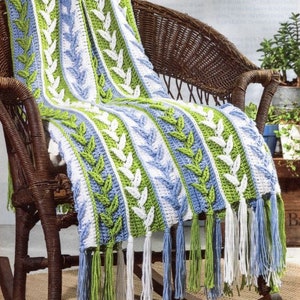 Könnte beinhalten: Eine handgehäkelte Decke mit einem Blattmuster in Weiß, Grün und Blau. Die Decke ist über einem Schaukelstuhl aus Korbgeflecht drapiert. Die Decke hat Fransen in passenden Farben.