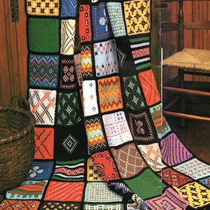 May include: A multi-colored crochet blanket featuring a grid of square patches with diverse geometric patterns in shades of red, blue, green, yellow, and white, all bordered by black yarn and finished with a black fringed edge at the bottom.