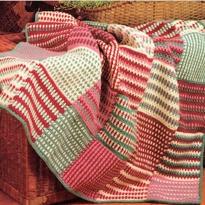 Op de afbeelding: Een gehaakt deken met een patchwork ontwerp in tinten roze, rood, groen en wit. Het deken is gemaakt met verschillende steekpatronen, waardoor een textuur ontstaat.