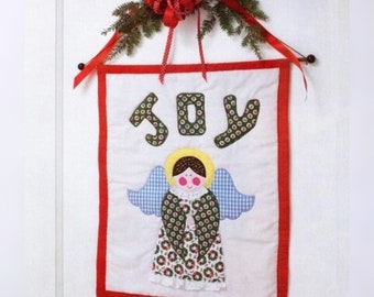 May include: A handmade Christmas wall hanging with the word "JOY" and an angel applique. The angel has blue gingham wings, a yellow halo, and a floral patterned dress. The banner is trimmed in red and adorned with a red bow and greenery.