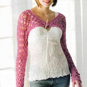 May include: A pink crochet shrug with long sleeves. The shrug is worn over a white top and blue jeans. The crochet design features an open, airy pattern. The top has a V-neck and a small decorative closure.