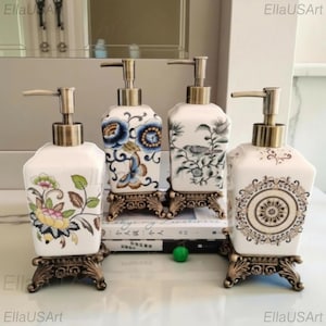May include: Four square ceramic soap dispensers with metallic pump heads and ornate bronze-toned bases. Each dispenser features distinct patterns: one with colorful flowers, one with blue flowers and a butterfly, one with a bird, and one with a circular mandala.