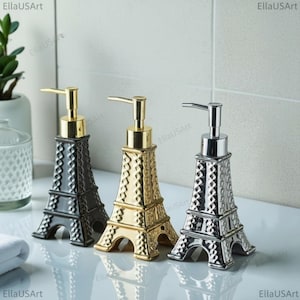 May include: Three soap dispensers shaped like the Eiffel Tower, featuring a black, a gold, and a silver metallic finish. Each dispenser has a textured lattice pattern on the exterior and a matching metallic pump mechanism at the top.
