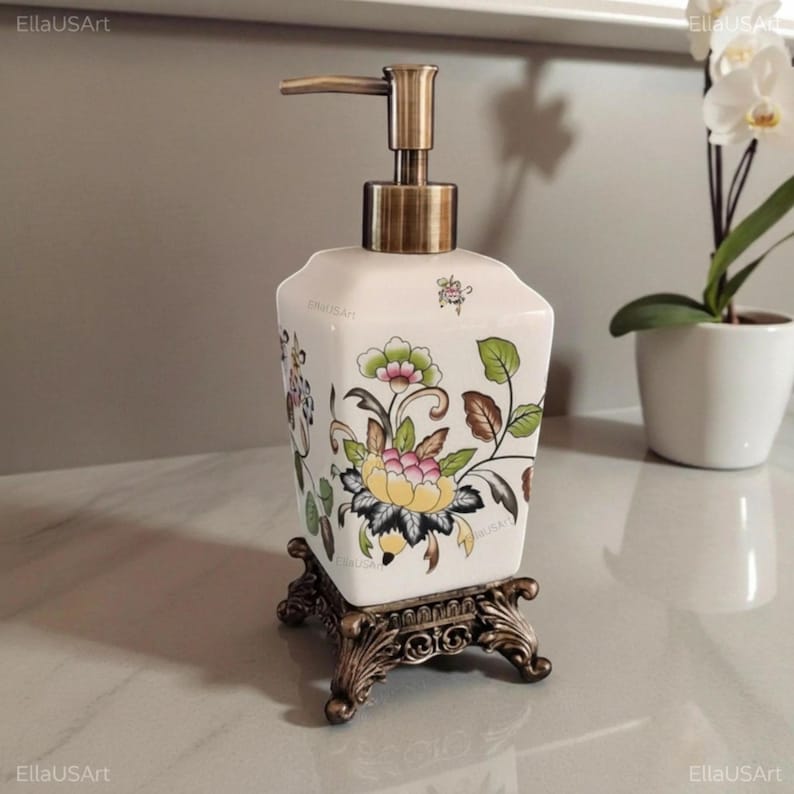 May include: A square white ceramic soap dispenser featuring a colorful floral pattern with yellow, pink, and green tones. The dispenser has a metallic bronze pump mechanism and rests on a decorative, ornate bronze-colored metal base with four feet.