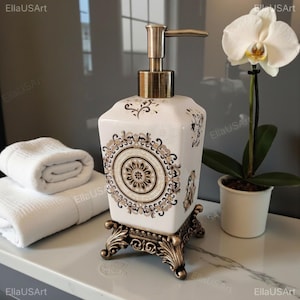 May include: A square white ceramic soap dispenser featuring a central circular mandala pattern in black, gold, and brown tones. The dispenser has a metallic pump and rests on an ornate, dark bronze-colored metal base with four decorative feet.