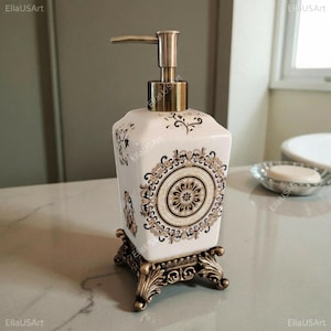 May include: A square white ceramic soap dispenser featuring intricate black and gold circular mandala-style patterns. The dispenser has a metallic bronze-toned pump mechanism and rests on a decorative bronze-toned base with ornate scrollwork feet.