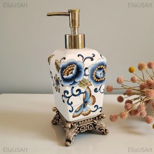 May include: A white ceramic soap dispenser with a square body featuring blue floral patterns and a butterfly motif. The dispenser has a metallic bronze-toned pump mechanism and rests on an ornate, four-legged bronze-toned metal base.