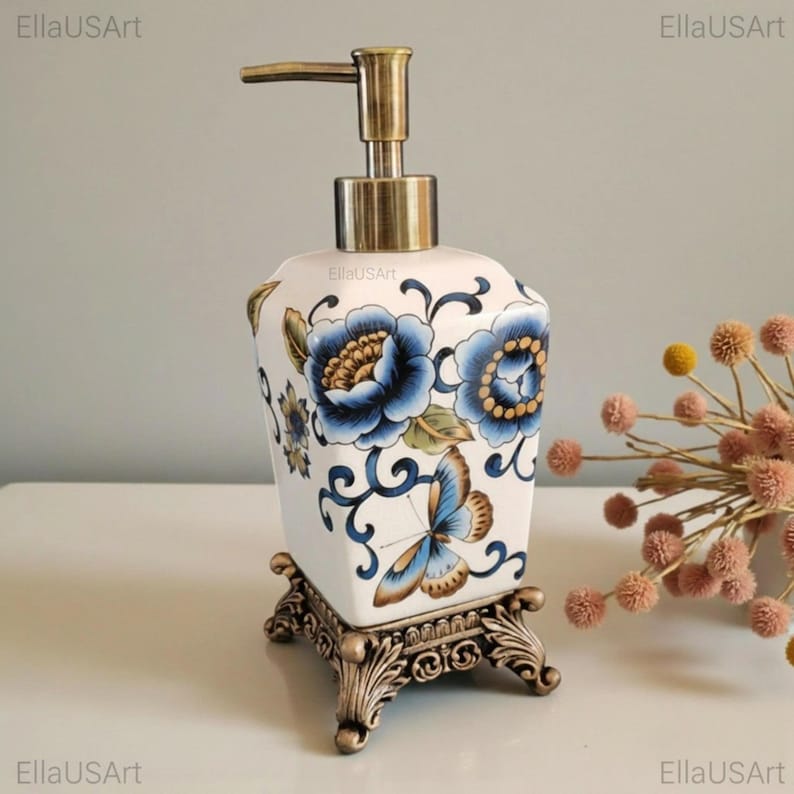 May include: A white ceramic soap dispenser with a square body featuring blue floral patterns and a butterfly motif. The dispenser has a metallic bronze-toned pump mechanism and rests on an ornate, four-legged bronze-toned metal base.