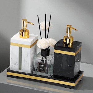 Op de afbeelding: Een decoratieve badkamerset met twee zeepdispensers met goudkleurige pompen, een glazen diffuser met stokjes en een dienblad. De dispensers hebben marmerachtige patronen in wit en zwart, geaccentueerd met gouden randen. De diffuser leest "AROMA Fresh Air".